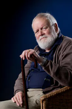 An elderly man with a white beard sat in a chair, holding a walking stick.
