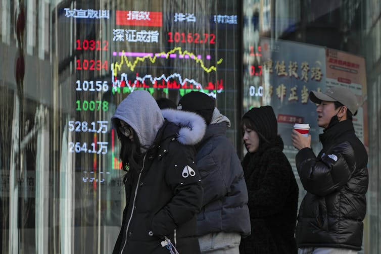 People walk past an electronic display board on a city street.