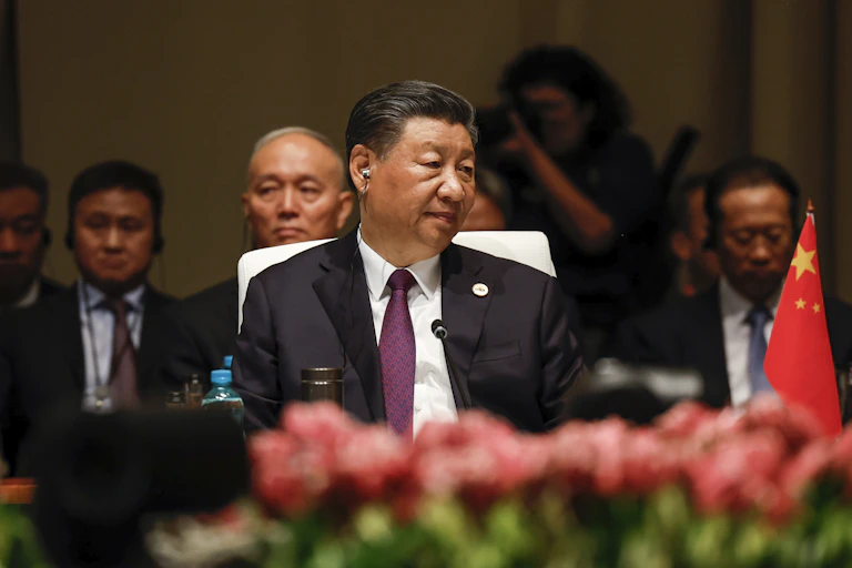 An Asian man sits at a conference with a small red flag next to him and fresh flowers in the foreground.