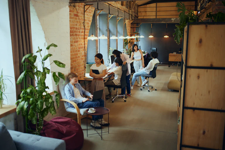 un espace de co working avec à gauche des femnêtres et des personnes jeunes qui travaillent.. le tout dans une ambiance qui évoque un ancien lieu industriel.
