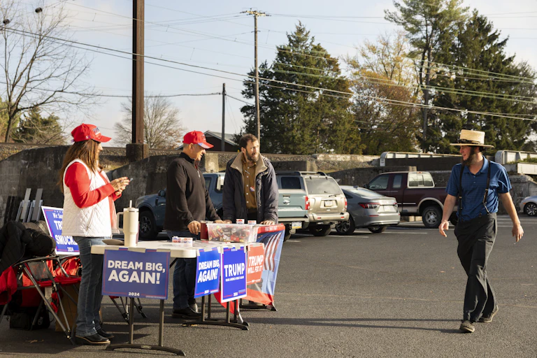 People stand behind a table adorned with signs supporting then presidential candidate Donald Trump as an Amish man walks by.