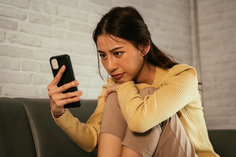 Une jeune femme, assise sur son canapé à la maison, regarde son téléphone portable.