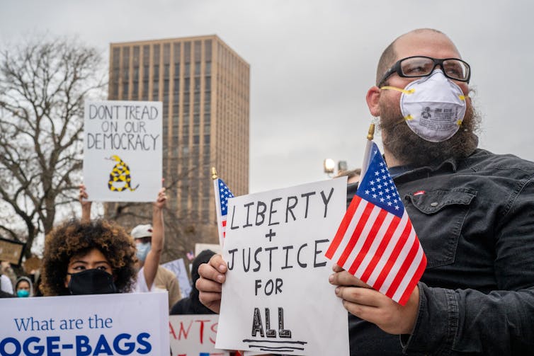 A man at a protest holding an American flag and a sign saying 'Liberty + Justice for ALL.'
