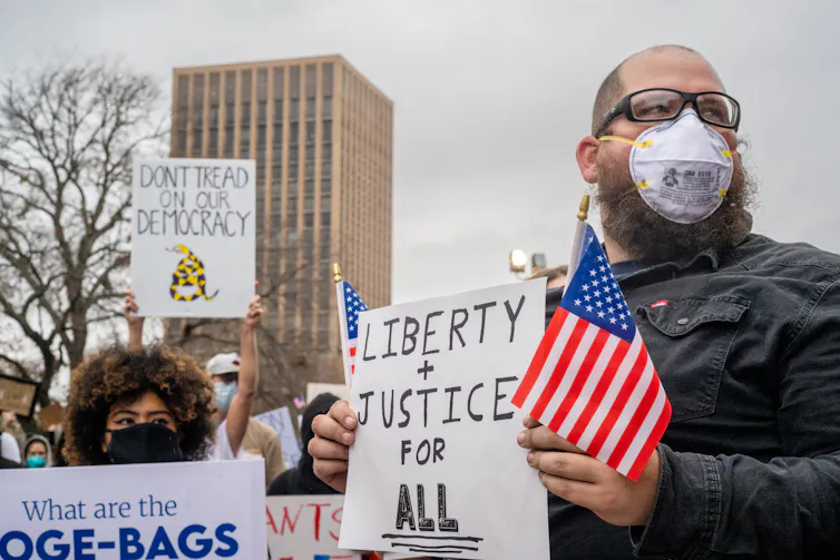A man at a protest holding an American flag and a sign saying 'Liberty + Justice for ALL.'
