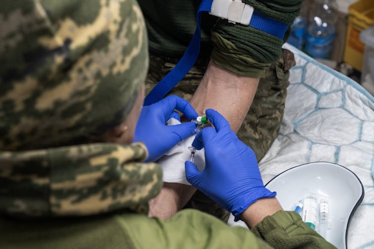 A medic in camo and blue sterile gloves places an IV into a soldier's arm.
