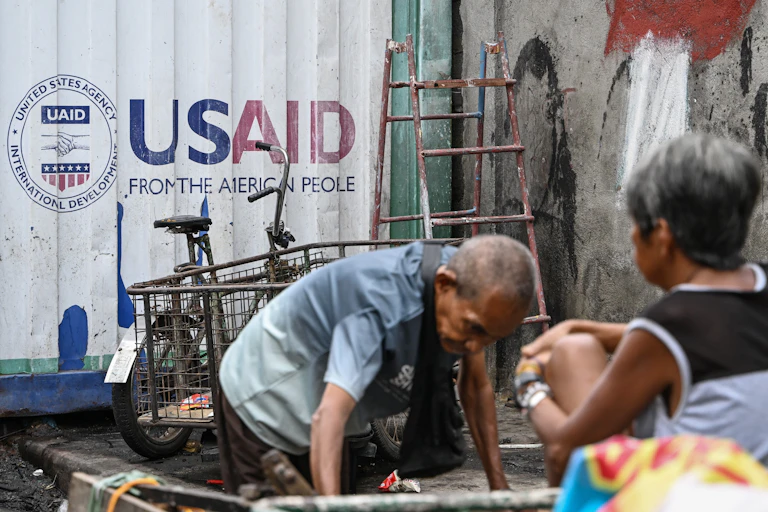 A man and woman rummage through various items near a cargo container with USAID signage.