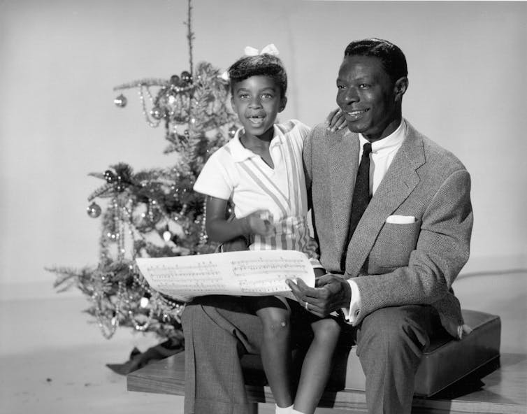 A photo of a young Black girl sitting on her father's lap, with a Christmas tree beside them.