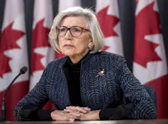 A woman with a grey bob and wearing glasses with a row of Canadian flags behind her.