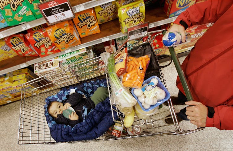 A baby resting in a shopping cart with cheesies, in a supermarket aisle of snack crackers