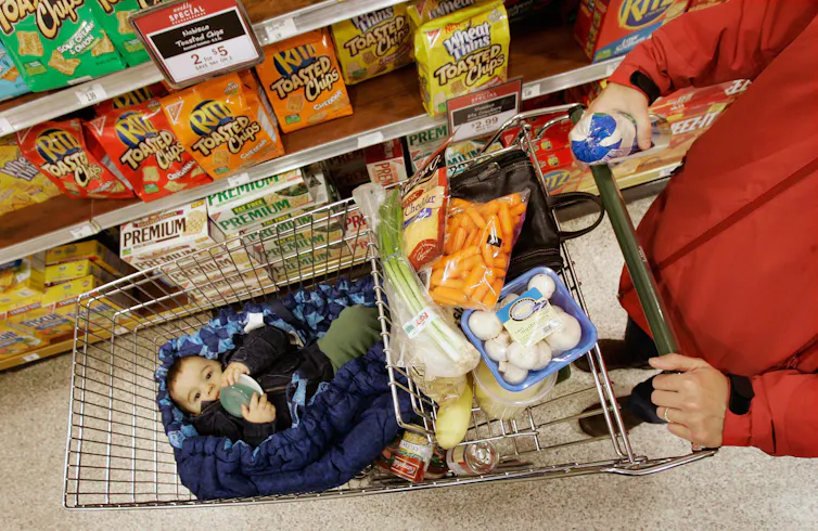 A baby resting in a shopping cart with cheesies, in a supermarket aisle of snack crackers