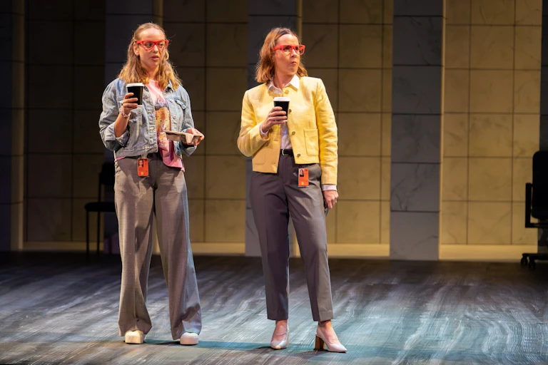 Two women stand on stage, drinking coffee.