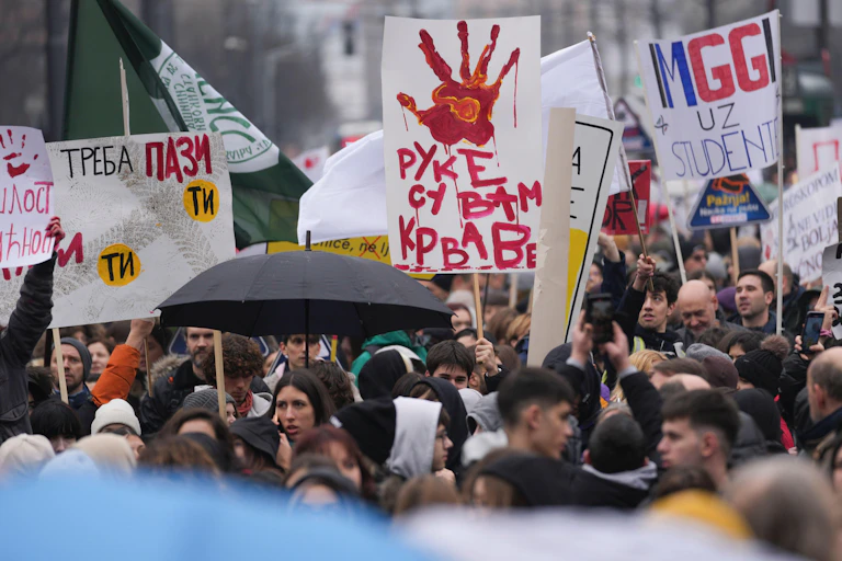 A crowd of protesters carrying signs in the Serbian language, one with a red handprint