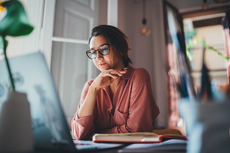 Woman sitting at a table in front of a laptop, her hand holding a pen and resting against her chin.