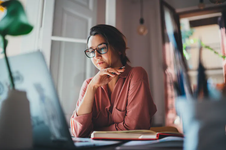 Woman sitting at a table in front of a laptop, her hand holding a pen and resting against her chin.