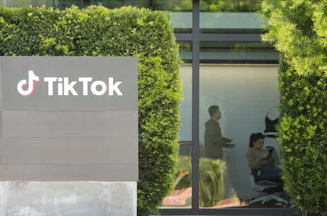 A sign says 'TikTok' on a gray wall in front of tall hedges and windows that show to people inside an office.