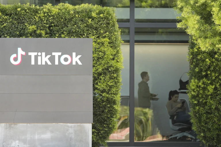 A sign says 'TikTok' on a gray wall in front of tall hedges and windows that show to people inside an office.