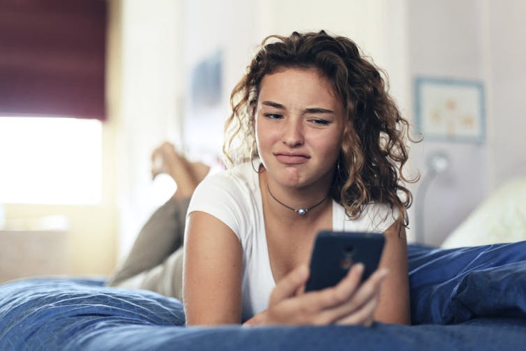 A young woman with curly hair rests on a bed with a blue cover, looking quizzically at her cellphone.