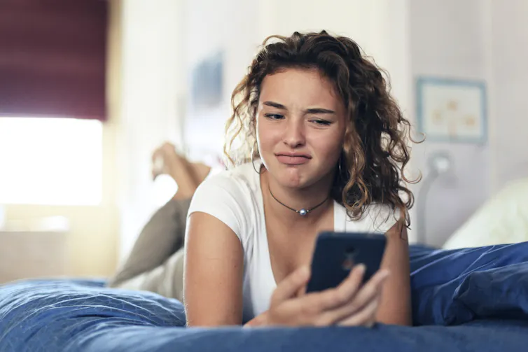 A young woman with curly hair rests on a bed with a blue cover, looking quizzically at her cellphone.