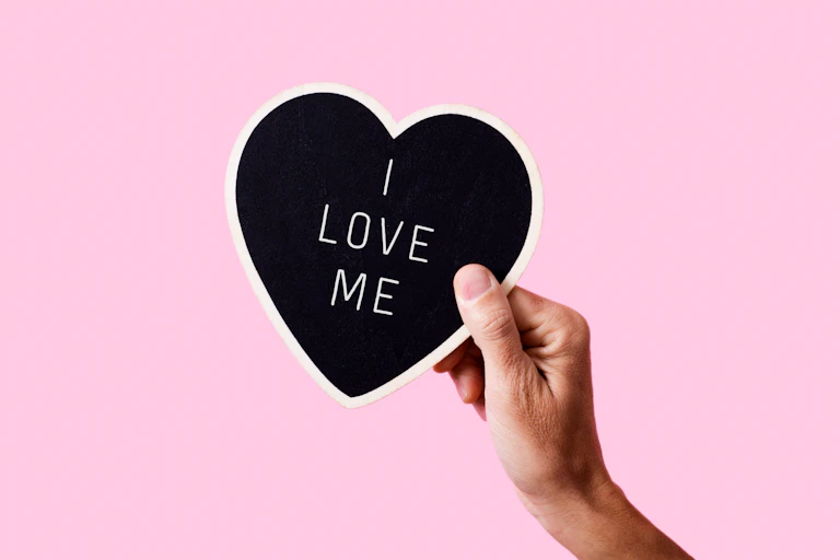 Hand holding a heart with the words "I love me" written on it.