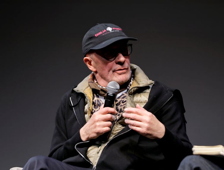 Older man wearing a hat and speaking into a microphone while seated.