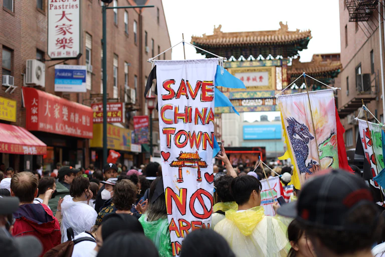 People gather in the streets of a Chinese neighborhood and hold signs, including one that says 'Save Chinatown - No Arena'