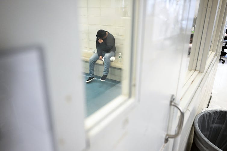 A man sits in a cell with head in hand, seen through a small window in the cell door