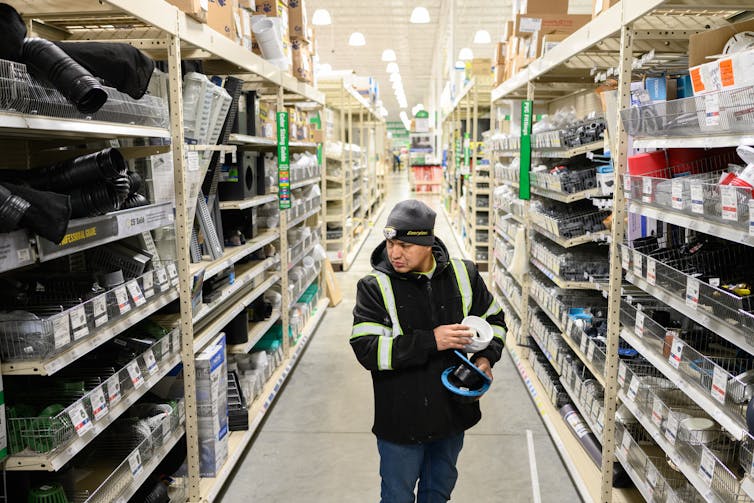 A man wearing a hat and a jacket holds items and walks down an empty aisle of what looks like a hardware store.