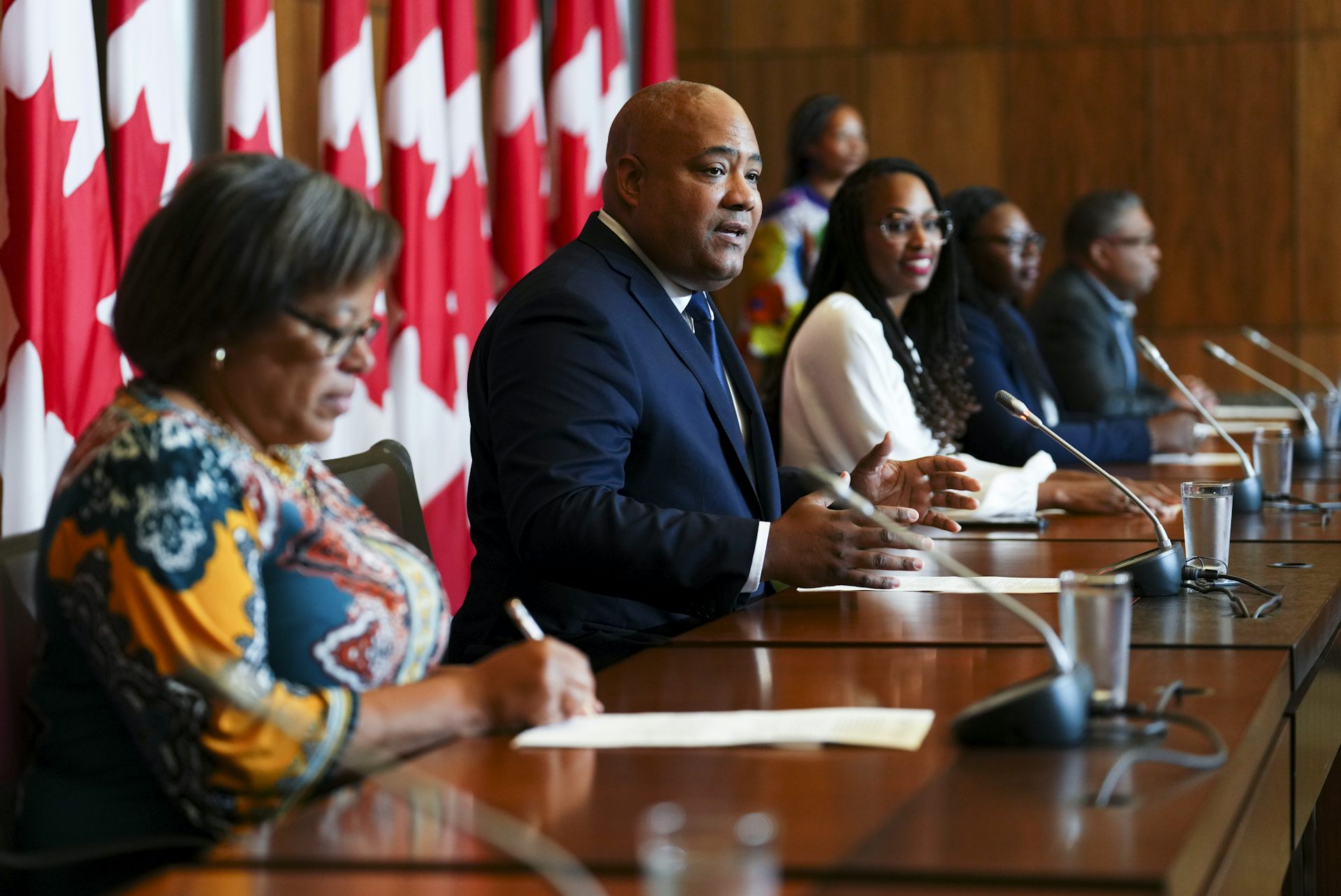 Black men and women sit in a row along a table with Canadian fags in the background. A man in a dark blue suit in the centre speaks into a mic
