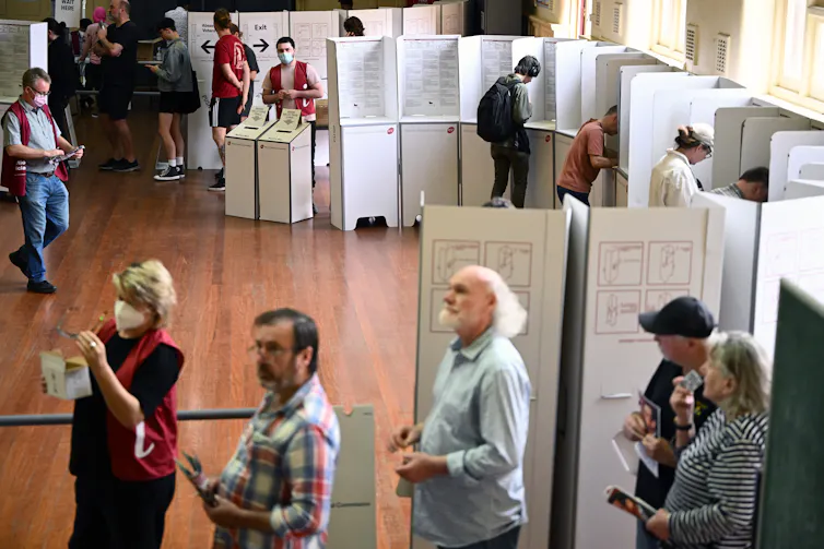Several men and women waiting in a room with poliched floors and cardboard voting booths