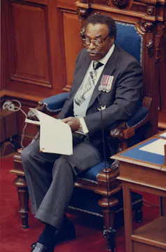 A black man in a dark suit sits on a wooden chair with blue cushions in a wood paneled room reading a speech