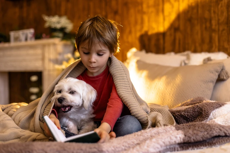 Un enfant assis sur un lit en train de lire avec son chien.