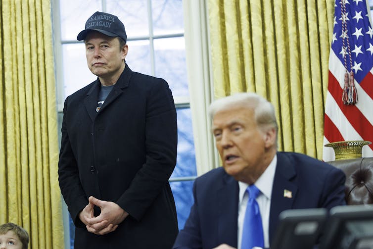 Donald Trump sitting at his desk in the oval office, while Elon Musk, wearing a black Make America Great Again hat, stands over his shoulder.