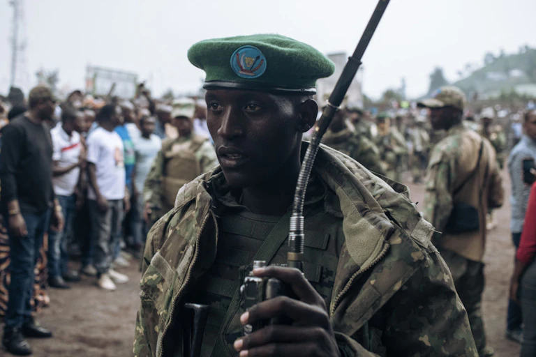 A man in army fatigues in the foreground with a crowd of people standing behind him.