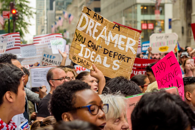 A crowd of people hold signs on a city street. The most prominent one reads "I am a DREAMER. You can't deport IDEALS. #DACA"