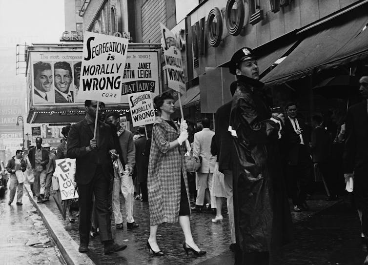 People in a black-and-white photo demonstrate outside a theater, holding signs that say 'segregation is morally wrong.'