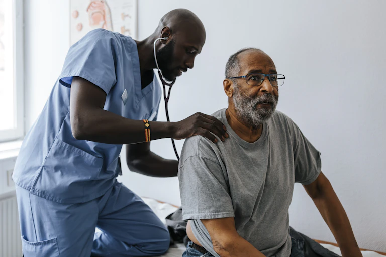 A man in medical scrubs kneels on an examination table as he checks another man's back with a stethoscope. There is a body diagram on the wall.