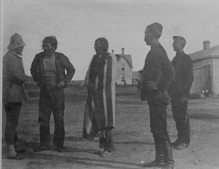 Man standing wrapped in a blanket with figures in military uniforms on two sides and one man in plain clothes.