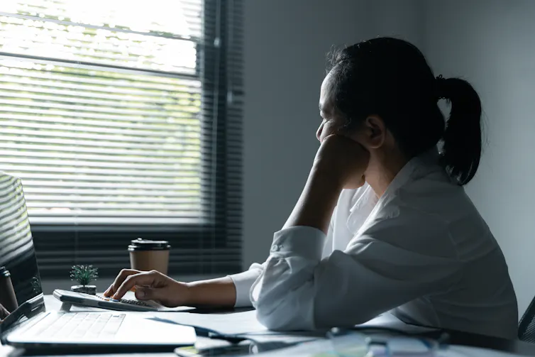 A woman looks pensively out the window of an office while seated at a desk