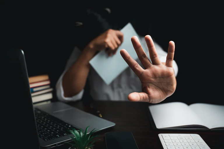 A person sitting at a desk ducks their head behind a piece of paper and puts out a hand toward the camera