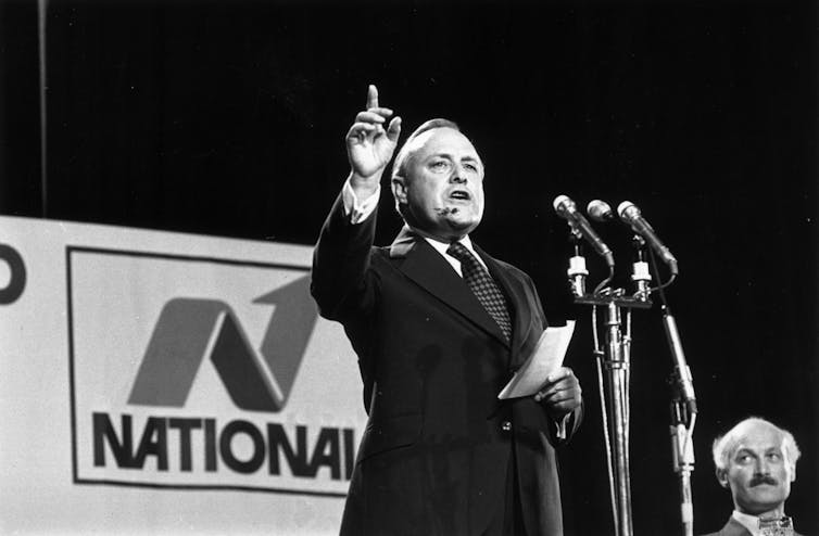 Black and white photo of older politician speaking into microphones with party banner behind him.
