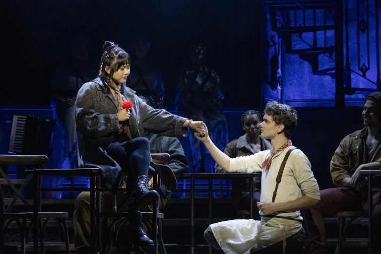 Production image: a woman sits on a table, holding a red flower. A man kneels on the ground, holding her hand.