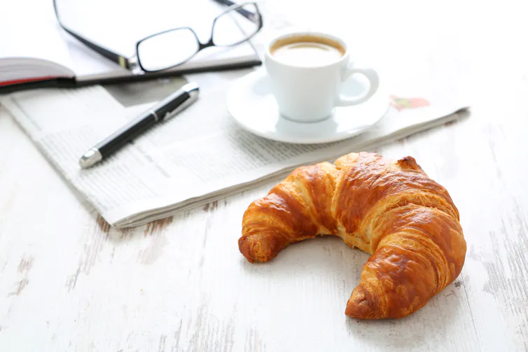 Glasses, a pen, a notebook on a newspaper, with a cup of coffee. A croissant sits on table too.