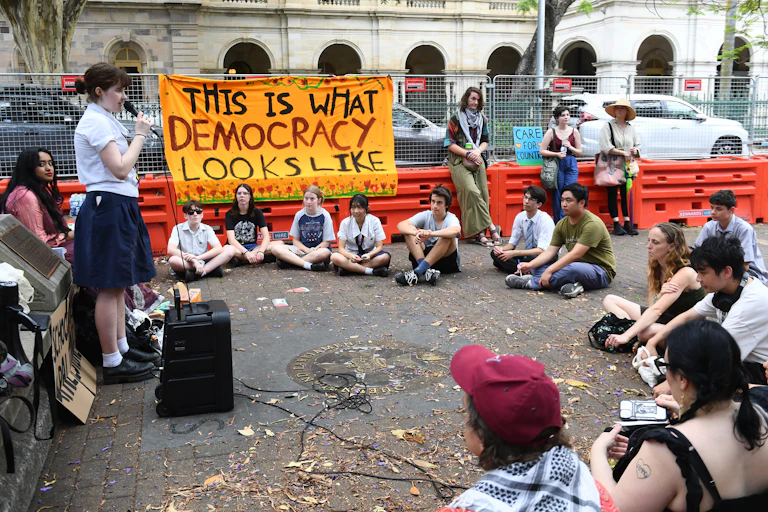 A school students stands with a microphone in front of a small circle of young people. A sign says 'this is what democracy looks like'.