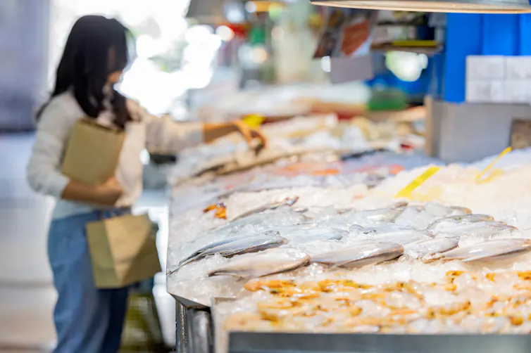 A woman carrying shopping bags selects some fresh seafood on ice at the supermarket.