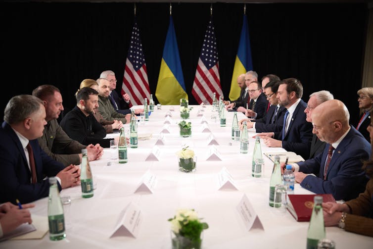 Volodymyr Zelensky and J.D. Vance and aides sit across a negotiating table from each other.
