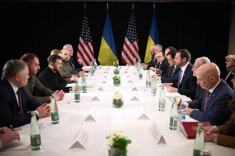 Volodymyr Zelensky and J.D. Vance and aides sit across a negotiating table from each other.