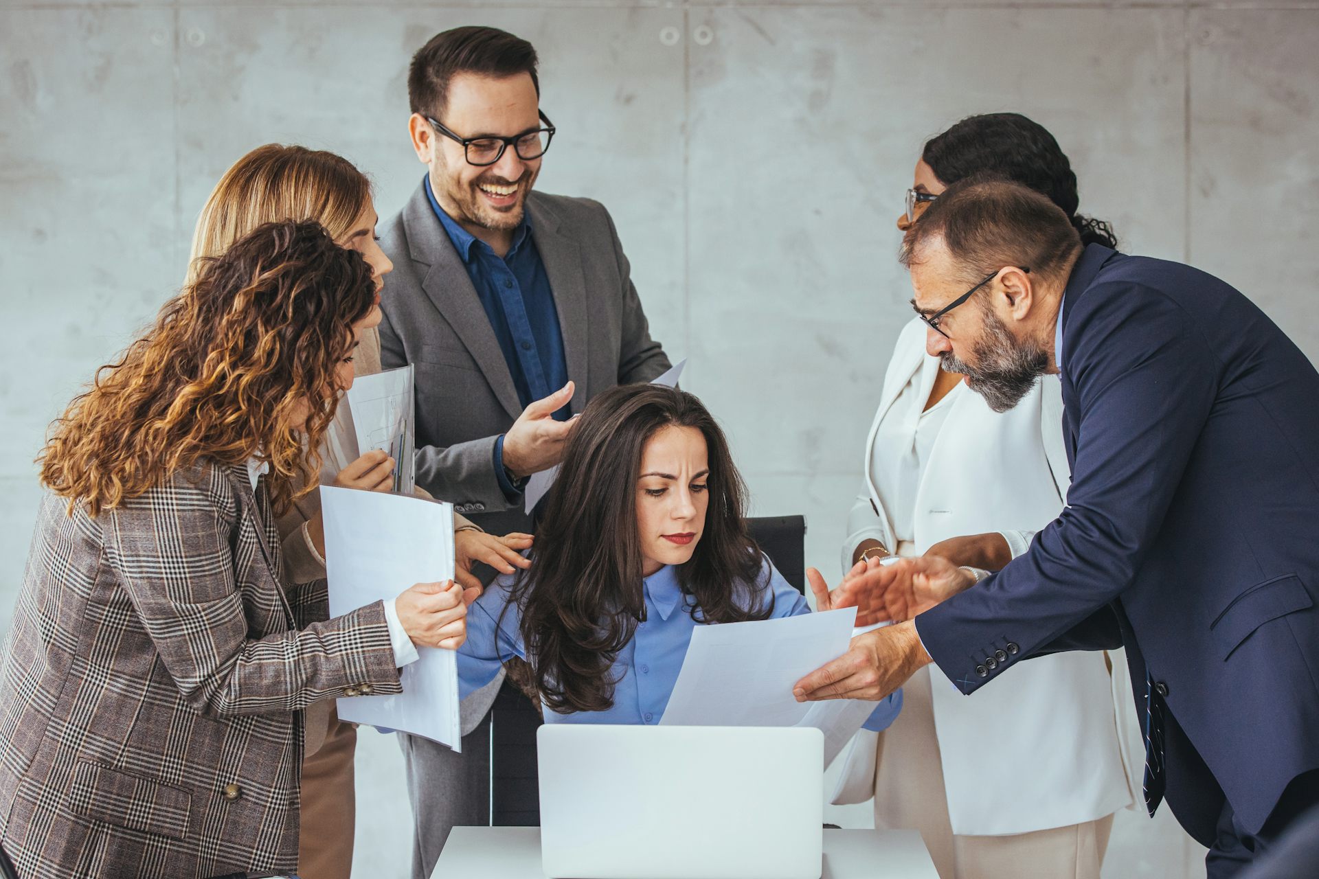 Five adults in business attire are surrounding a colleague who is seated and reading a document.