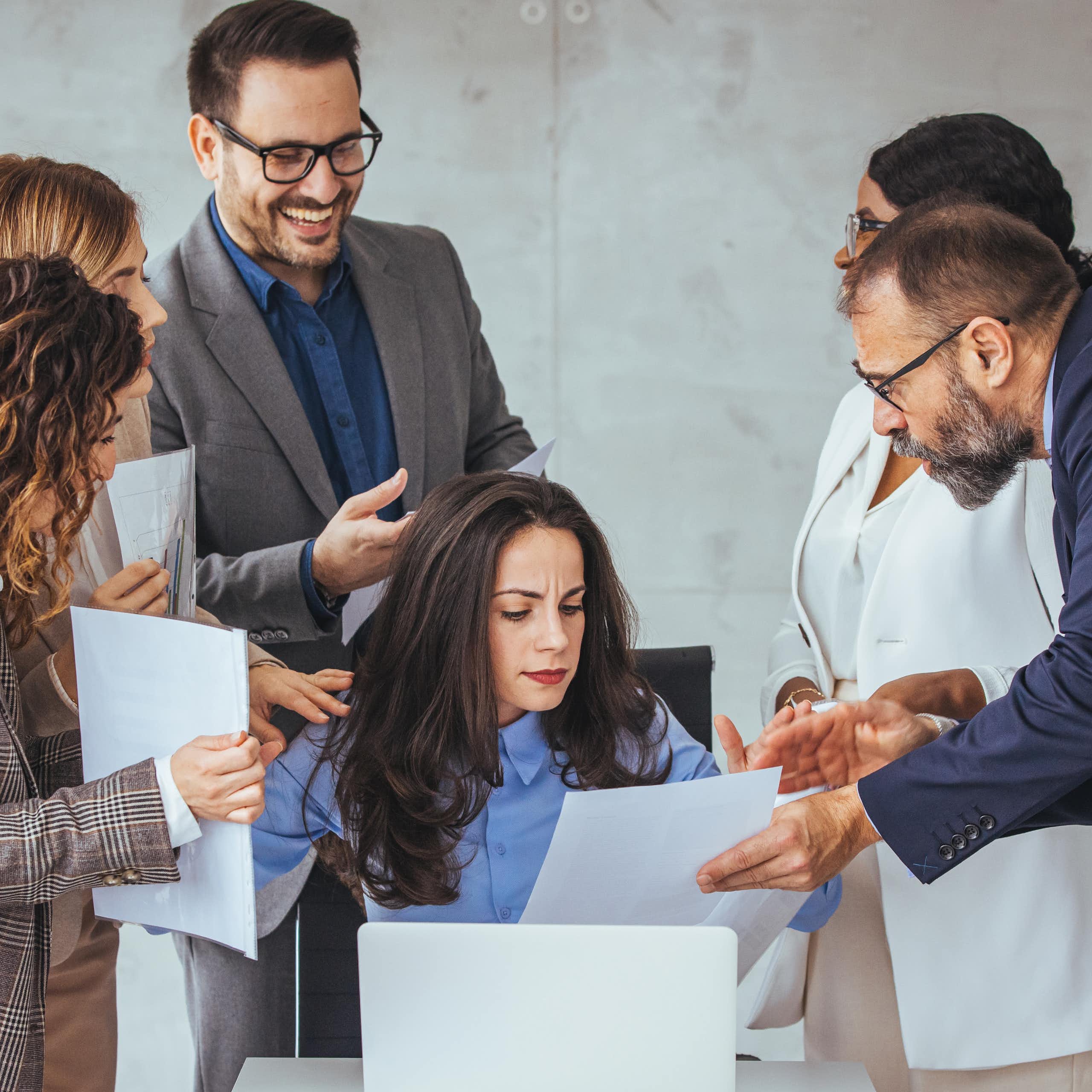 Five adults in business attire are surrounding a colleague who is seated and reading a document.