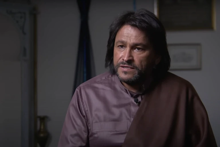 A man in a purple robe with long dark hair sits talking intently.