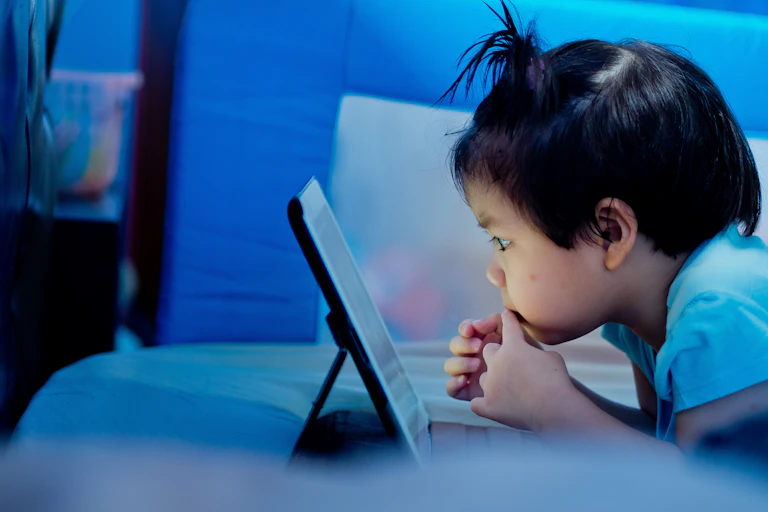 Young child lying on a bed watching a computer tablet up close.